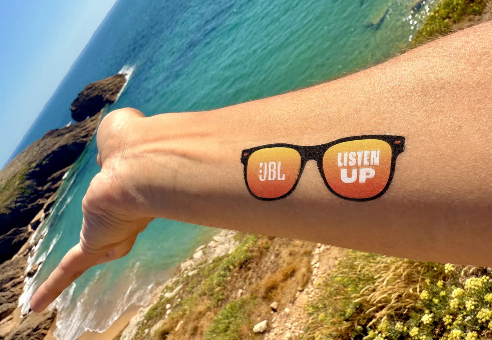 JBL "Listen Up" sunglasses branded tattoo on a forearm at the beach